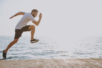 Side view full length portrait of handsome young bearded athletic man guy 20s in casual white t-shirt black shorts posing training running jumping looking aside at sunrise over the sea outdoors.