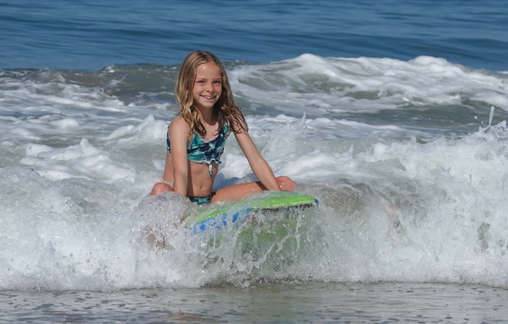 A young blond girl smiles broadly as she rides a wave sitting on a boogie board 