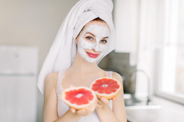 Pretty young girl in bath towel and facial mask with grapefruit slices, showing to camera....