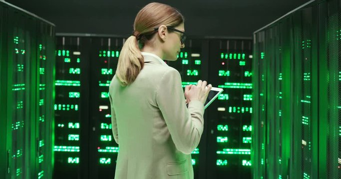 Rear on Caucasian young woman in glasses walking at servers with tablet device, tapping and checking big data. Back view on female analytic in data storage working on digital security of database.
