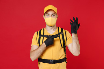Delivery employee man in yellow cap face mask gloves t-shirt uniform thermal bag backpack with food work courier service during quarantine coronavirus covid-19 virus isolated on red background studio.