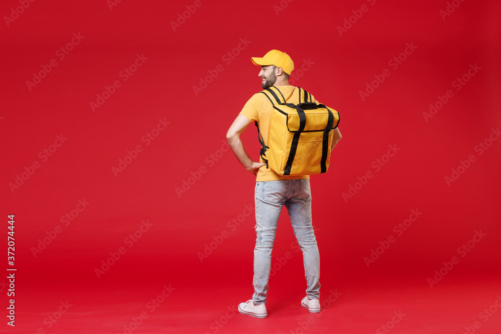 Wall mural Full length back view delivery employee man guy male 20s in yellow cap t-shirt uniform thermal food bag backpack work courier service in quarantine covid-19, standing isolated on red background studio