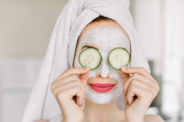 Close up of beautiful young woman with facial mask on her face holding slices of fresh cucumber covering her eyes, on home interior background. Skin care and treatment, spa at home