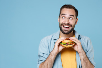 Excited young bearded man guy 20s wearing casual clothes posing holding in hands american classic fast food burger looking aside up isolated on pastel blue color wall background studio portrait.