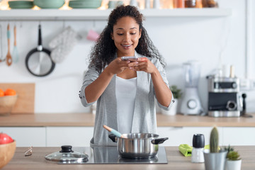 Happy young african american woman cooking healthy food while taking a photo with her mobile phone in the kitchen at home.
