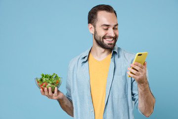Cheerful smiling young bearded man guy 20s wearing casual clothes posing holding vegetable salad in glass bowl using mobile cell phone isolated on pastel blue color wall background studio portrait.