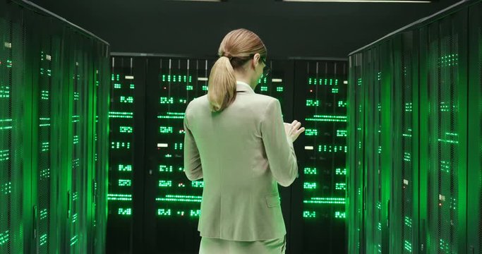 Rear of Caucasian young woman with walking among servers and checking work of big data processors. Back view Female analytic in data storage strolling in information security office with tablet device