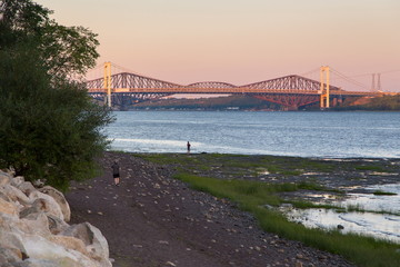 High angle back view of men strolling or standing on a beach during a summer golden hour evening, with the Pierre-Laporte and Quebec bridges in the background, Cap-Rouge area, Quebec City, Canada 
