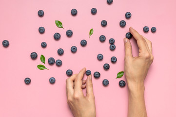 Fresh juicy blueberries with green leaves and female hands on pink background. Blueberries background. Flat lay top view. Healthy berry, organic food, antioxidant vitamin blue food. Blueberry pattern