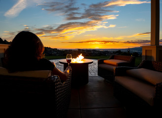 A woman relaxes with a glass of wine at night in front of an outdoor firepit on a patio of a luxury...