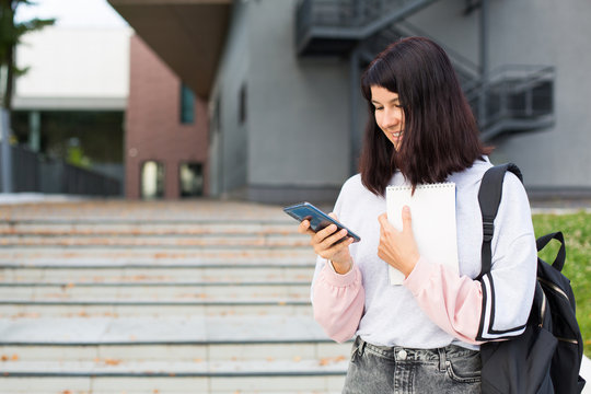 A female student with a black backpack holds a smartphone and Notepad on the threshold of the University. Higher education, the beginning of the school year, return to school, gadgets. Copyspace