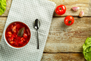 A plate of borscht with chicken on a napkin with a spoon on a wooden table next to garlic and...