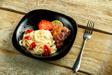 Black plate of spaghetti with baked chicken with a fork on a wooden table.