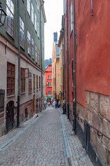 View of a narrow street in the historic part of an ancient European city, with historic buildings, interesting architecture, residents and tourists, one cloudy, cold summer day