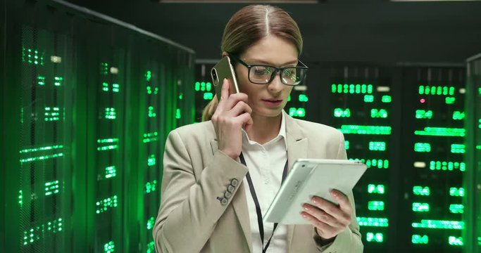Caucasian beautiful woman among servers with tablet computer and talking on mobile phone. Pretty female analytic in data storage working on digital security, speaking on cellphone with gadget in hand.