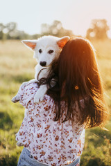 Fototapeta premium Stylish young woman hugging cute white puppy in warm sunset light in summer meadow. Happy girl holding adorable fluffy puppy. Beautiful atmospheric moment. Adoption concept.