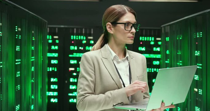 Caucasian young woman in glasses standing at servers with laptop computer, typing on keyboard and checking big data processor. Female analytic in data storage working on digital information security.