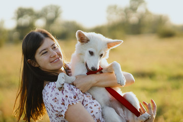 Stylish young woman hugging cute white puppy in warm sunset light in summer meadow. Happy girl...