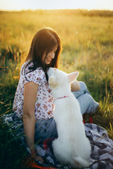 Summer travel with pet. Happy woman sitting with cute white puppy in summer meadow in sunset warm light. Stylish girl relaxing with her adorable fluffy puppy on a picnic.