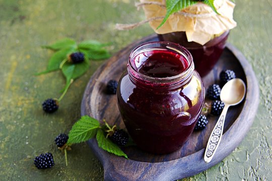 Blackberry and red currant jam in a curly glass jar on a green concrete background.