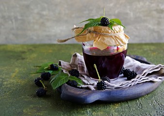 Blackberry and red currant jam in a curly glass jar on a green concrete background.
