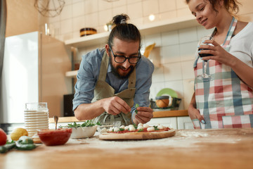 Best cook. Young couple making pizza together at home. Man in apron, professional cook adding basil on the dough while woman drinking wine. Hobby, lifestyle