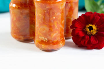 fermented tomato, paprika, aubergine, onion  sauce in glass jars on  white wooden background and red flower