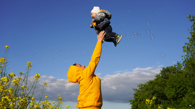 Dad Plays With A Small Child. Father Throws The Baby Into The Air On A Sunny Day Against The Background Of Blue Sky, Yellow Flowers And Soap Bubbles. Carefree Childhood, Parental Love, Parenting