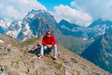 Naklejka na ściany i meble Young man in sunglasses sitting on mountain in sunny weather. Adult male in red hoodie with hood enjoying beautiful view in mountainous area.