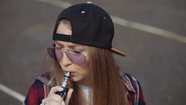 Close up pretty young hipster woman in black cap vape ecig. Girl vape popular ecig gadget, vaping device.Happy brunette vaper girl with e-cig. Portrait of smoker female model with electronic cigarette