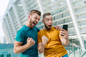 Men celebrating money win in online sport gambling application with football stadium on the background - obrazy, fototapety, plakaty