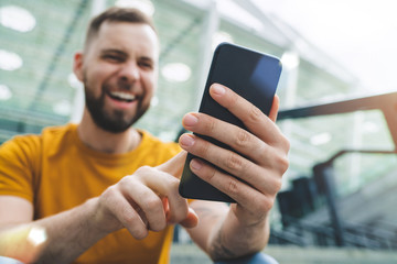 Young handsome smiling bearded man sitting at stadium steps and making bets online on his smartphone using mobile bookmaker application
