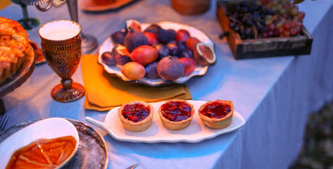 Closeup pastry - berry tart on white plate, figs and grapes. Autumn evening photo shoot, romantic dinner outdoors. Table with tablecloth and decoration - wine glasses, napkin, table setting