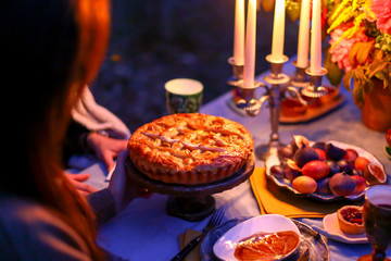 Autumn evening photo shoot. Young woman cut cake with knife. Romantic dinner at twilight nature outdoors. Table decoration closeup - figs, plates, candelabra with candles, flowers. Red dahlia bouquet