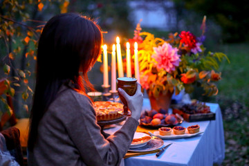Autumn evening photo shoot: young woman with wine glass at romantic dinner in twilight nature outdoors. Table decoration - cake, figs, plates, candelabra with candles, fall flowers. Red dahlia bouquet
