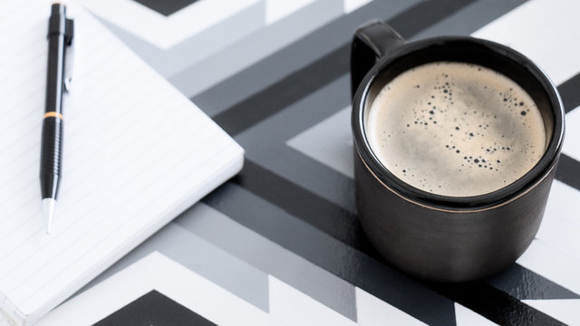 Vista cenital de taza de cer&aacute;mica negra de caf&eacute; sobre bandeja con estilo moderno junto a anotador de hojas rayadas y lapicera negra en un ambiente iluminado y blanco