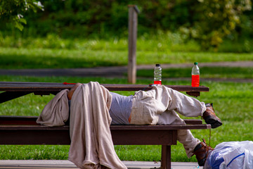 A homeless caucasian man is sleeping on a picnic table in a shaded area of a park. He is covering his head with a dirty blanket. His belongings are in plastic bags and he has some drinks on table.