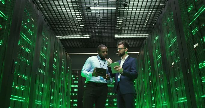 Mixed-races men with tablets devices. Software cryptographers walking at servers and checking big data information. Multiethnic males analytics coworkers discussing digital security. Analyzing data.