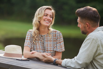 Portrait of romantic couple holding hands and looking at each other with love while sitting at outdoor table by lake, copy space