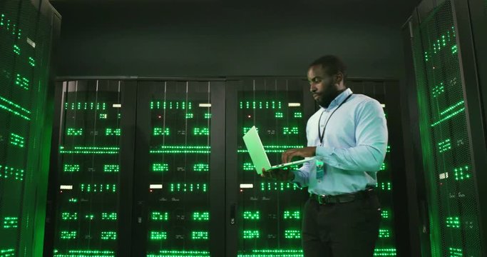 African American young man walking among servers with laptop computer and checking work of big data processors. Male analytic in data storage browsing and typing on keyboard.