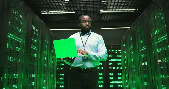 African American young man walking among servers with laptop computer and checking work of big data processors. Male analytic in data storage working on information security.