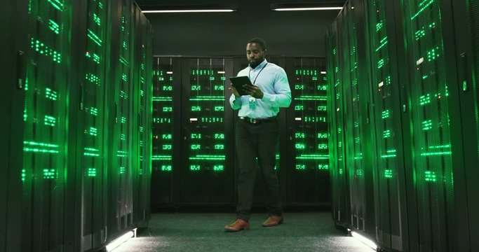 African American young man walking among servers with tablet device, tapping on screen and checking big data processors. Male analytic in data storage working on digital information security.