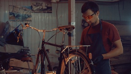 theme small business bike repair. A young Caucasian brunette man wearing safety goggles, gloves and an apron uses a hand tool to repair and adjust the bike in the workshop garage