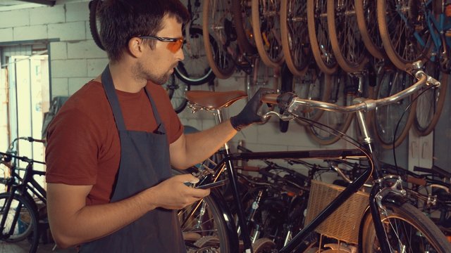 theme small business bike repair. Young caucasian brunette man wearing safety goggles, gloves and fartukhe uses mobile phone technology, takes notes, checklist in bicycle workshop