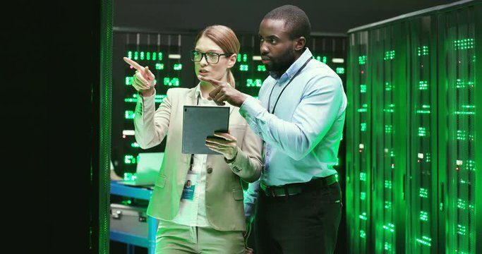Multiethnic man and woman talking and checking database servers. Using tablet device. Couple of mixed-races coworkers discussing security. Male and female analytics cryptographers in data center.
