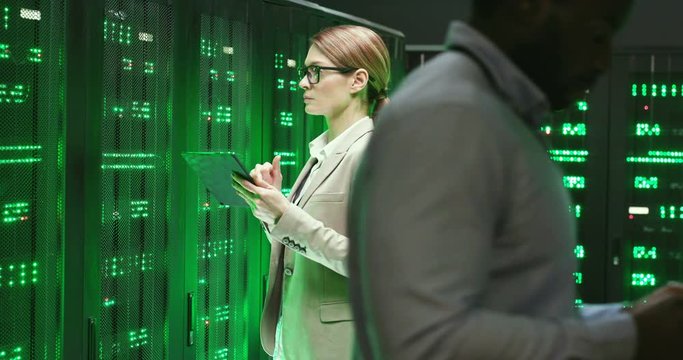 Caucasian woman, cryptographer standing at server with tablet device and checking big data processors. Female analytic working on digital security. Males colleagues with laptop and tablet passing by.