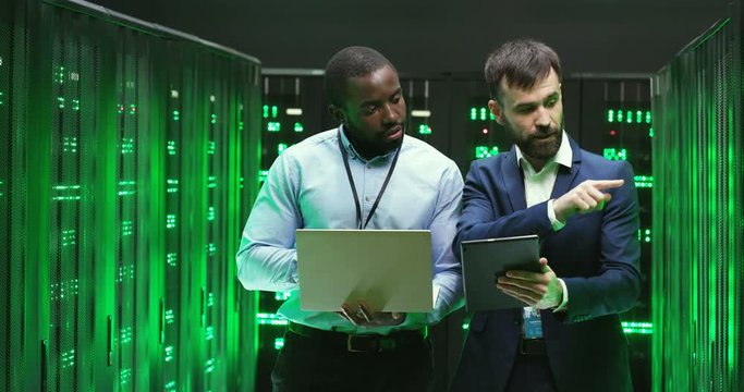 Multiethnic men talking and checking database storage with servers. Using tablet device and laptop computer. Mixed-races colleagues discussing security. Males analytics cryptographers.
