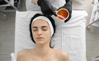 a professional beautician in his office picks up an orange cream mask to apply to a client who lies...