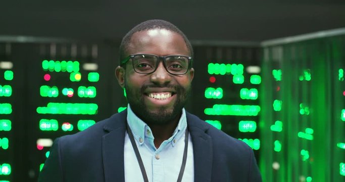 Portrait of handsome Caucasian young man in glasses looking at camera in big data storage room with many processors and smiling. Male system administrator standing among servers in datacenter.