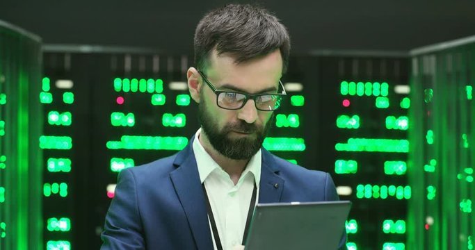 Close up of handsome Caucasian young man in glasses tapping and scrolling on tablet device in big data storage room with many processors. Male system administrator using tablet computer at servers.